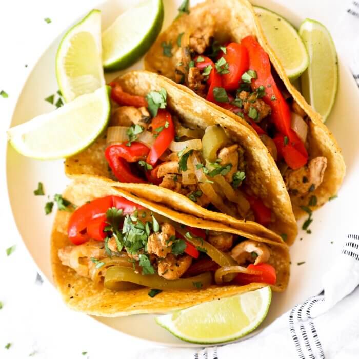 Overhead image of 3 taco shells filled with chicken fajitas. Topped with fresh chopped cilantro and lime wedges holding the tacos up.