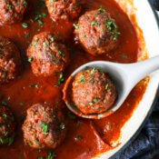 Overhead image of meatballs simmering in a skillet with marinara sauce and fresh parsley sprinkled on top. A serving spoon is scooping one meatball.