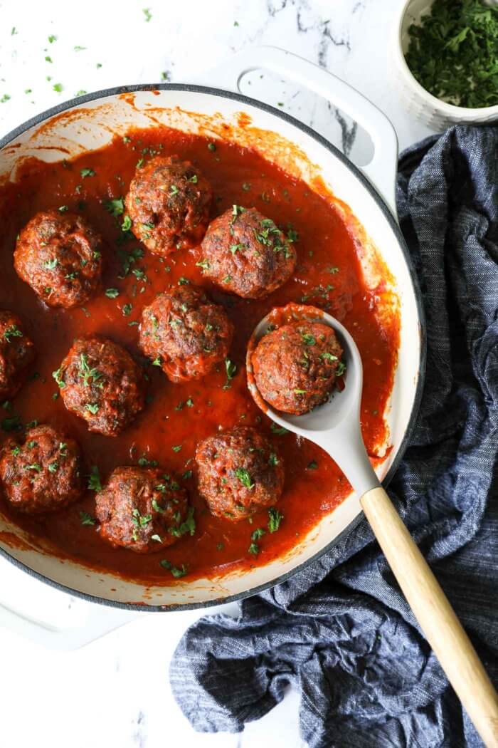 Pulled out overhead image of meatballs simmering in a skillet with marinara sauce and fresh parsley sprinkled on top. A serving spoon is scooping one meatball.