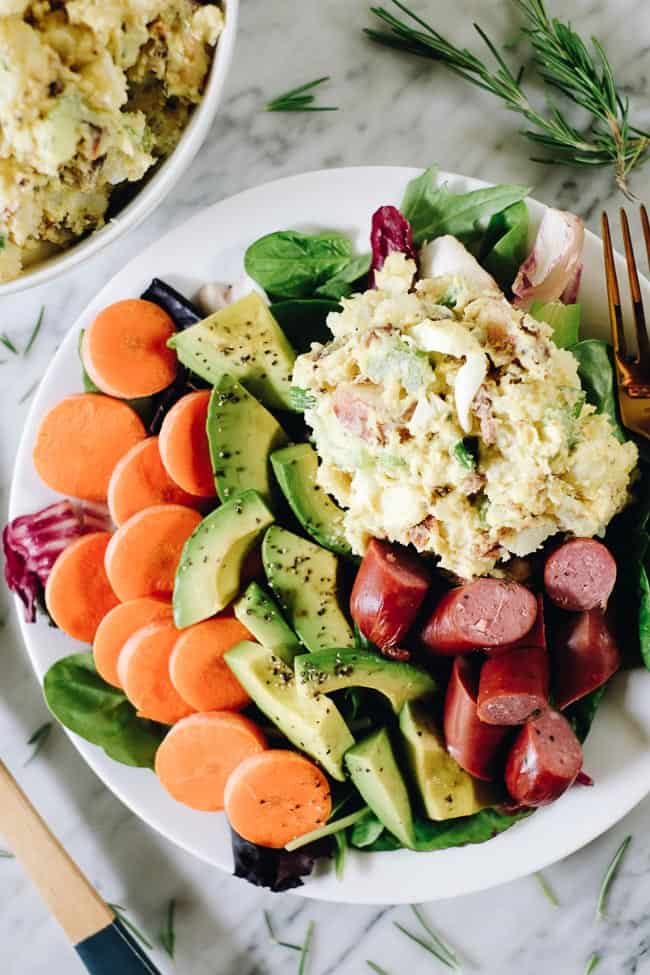 The best potato salad with bacon on a plate with greens, carrots, avocado and sausage. 