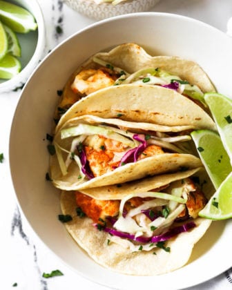 Flatlay image of three fish tacos situated in a bowl with a few lime wedges and chopped cilantro on top.