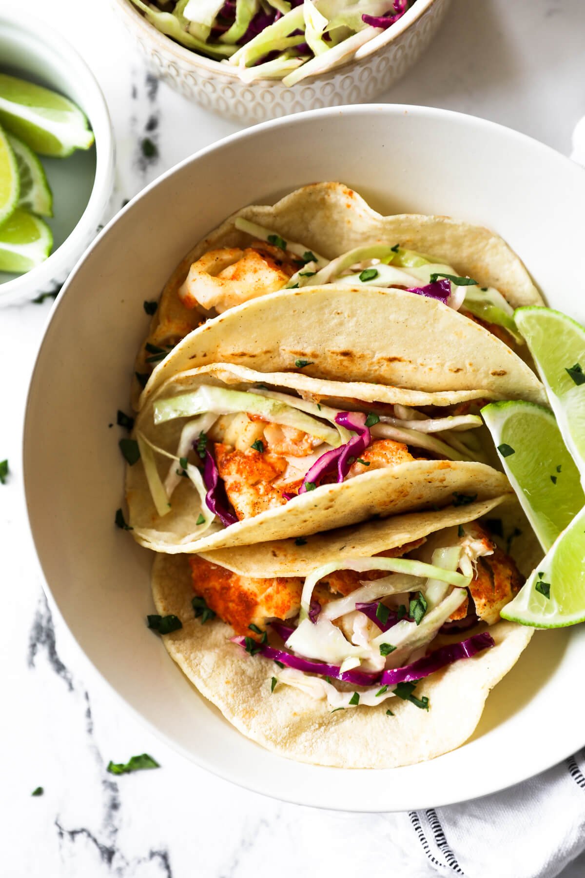 Flatlay image of three fish tacos situated in a bowl with a few lime wedges and chopped cilantro on top. 