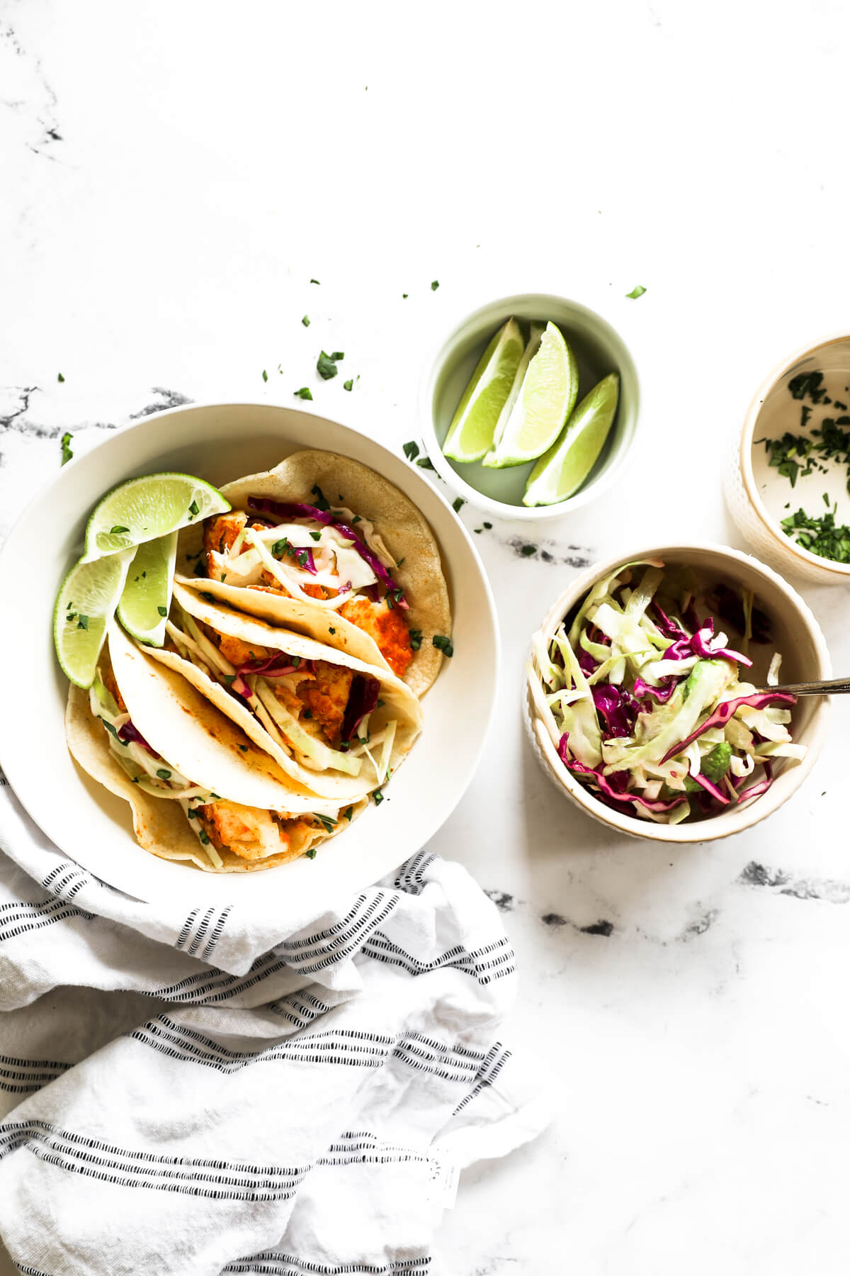Overhead image of a bowl with three fish tacos and smaller bowls with lime wedges, a citrusy coleslaw and chopped cilantro all on the side.