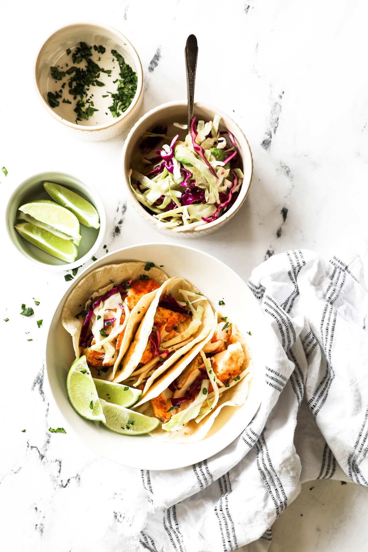 Overhead image of a bowl with three fish tacos and smaller bowls with lime wedges, a citrusy coleslaw and chopped cilantro all on the side.
