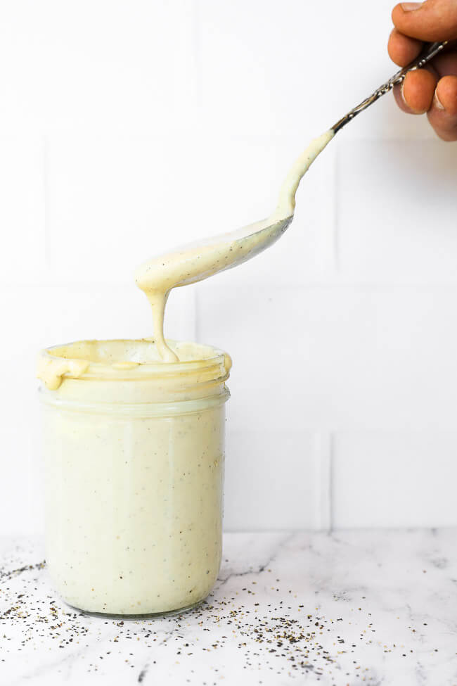 Vertical straight on image of jar of vegan alfredo sauce with a spoonful being pulled out of jar and dripping off spoon. 