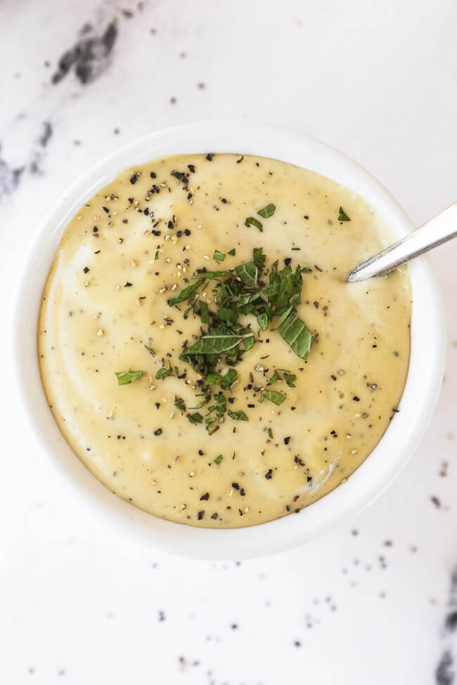 Vertical overhead close up of one ramekin filled dairy free alfredo sauce, topped with chopped lemon balm and cracked pepper with a spoon dug in. 