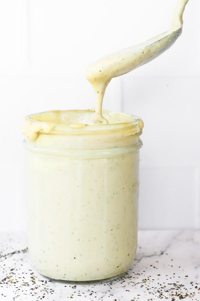 Vertical straight on image of jar of vegan alfredo sauce with a spoonful being pulled out of jar and dripping off spoon.