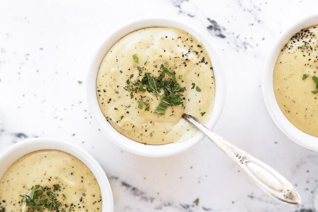 Overhead horizontal image of vegan alfredo sauce in ramekins with chopped lemon balm and cracked pepper on top. A spoon is dug into one ramekin. 