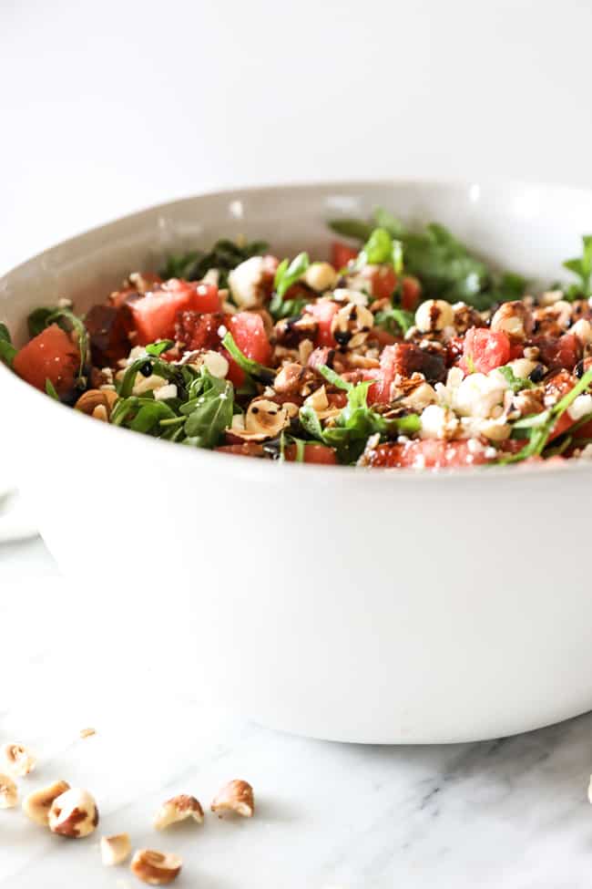 Angled vertical image of watermelon salad in a large serving bowl. Watermelon, arugula, hazelnuts, balsamic vinegar and optional feta cheese. 