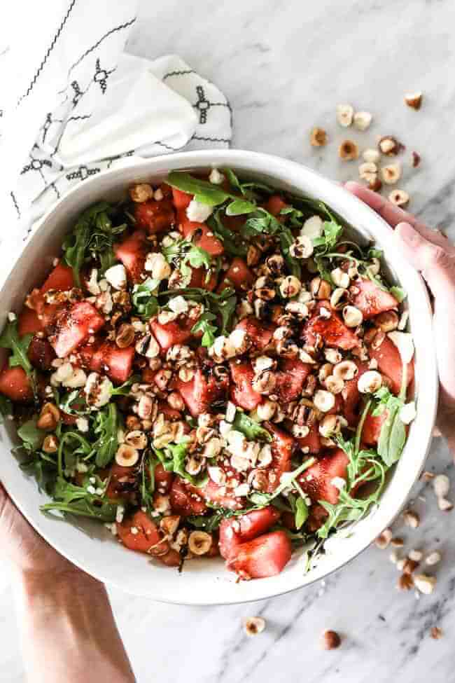 Overhead vertical image of holding watermelon salad in a large serving bowl. Watermelon, arugula, hazelnuts, balsamic vinegar and optional feta cheese. 