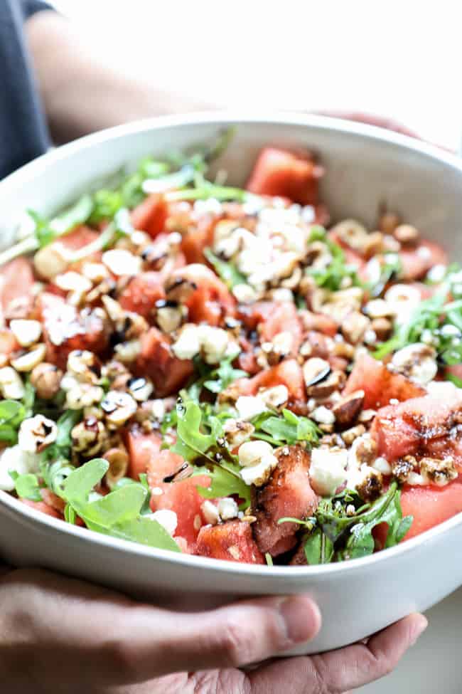 Angled vertical image of holding a large serving bowl of watermelon salad. Watermelon, arugula, hazelnuts, balsamic vinegar and optional feta cheese. 