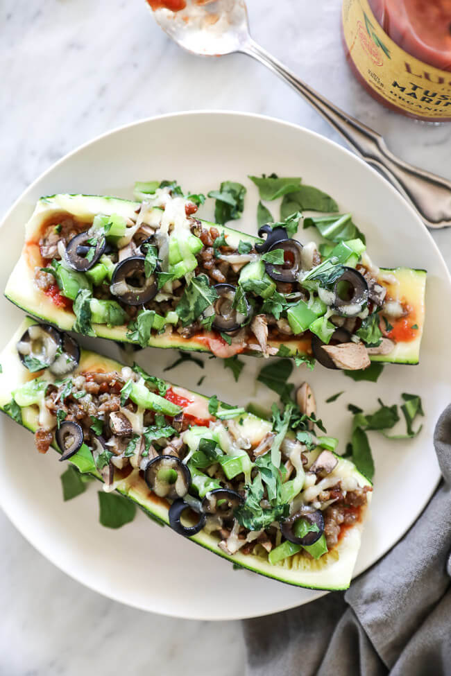 Overhead vertical image of zucchini pizza boats on plate. Loaded with marinara sauce, italian sausage, bell pepper, mushrooms, olives and cheese. Fresh basil sprinkled on top. 