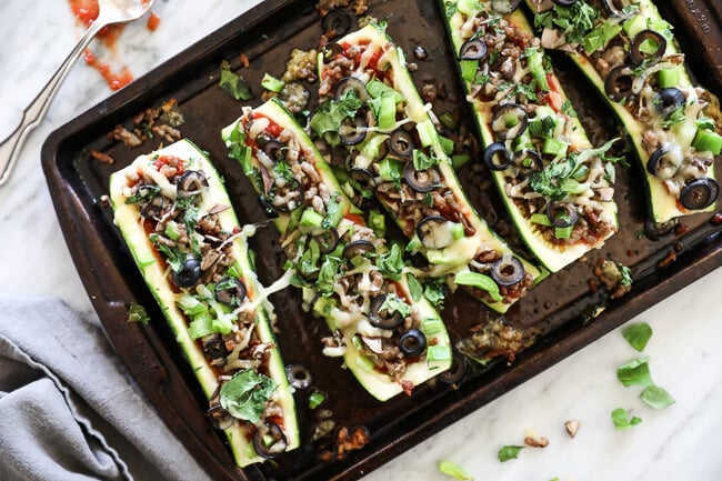 Overhead horizontal image of zucchini pizza boats on sheet pan. Loaded with marinara sauce, italian sausage, bell pepper, mushrooms, olives and cheese. Fresh basil sprinkled on top. 