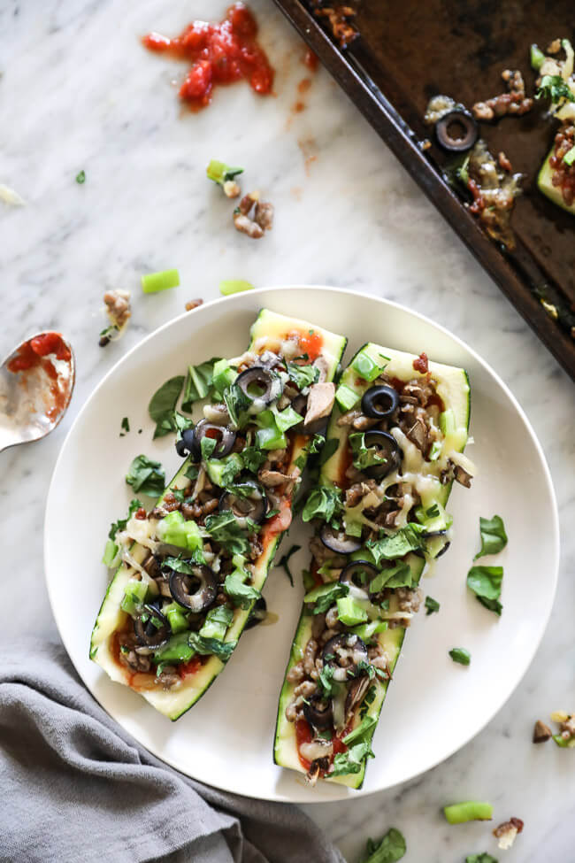 Overhead vertical image of zucchini pizza boats on sheet pan. Loaded with marinara sauce, italian sausage, bell pepper, mushrooms, olives and cheese. Fresh basil sprinkled on top. 